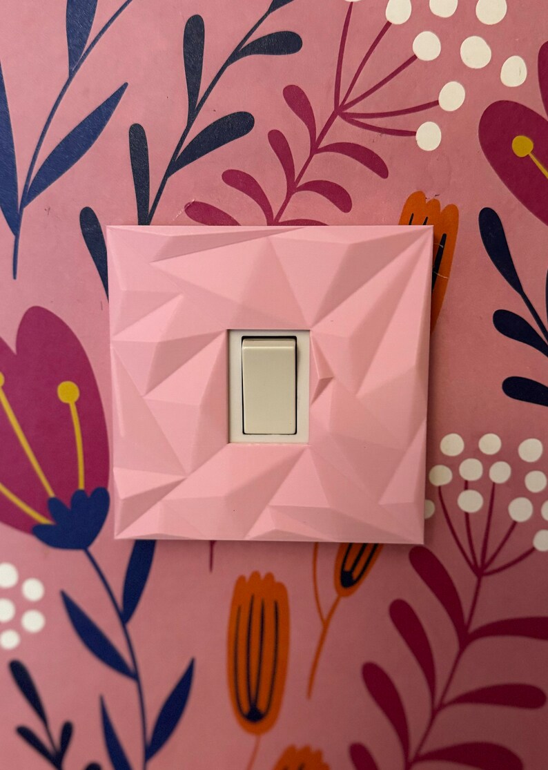 May include: A square light switch cover with a faceted, geometric pink surface texture. The cover features a central rectangular opening that reveals a standard off-white toggle switch mechanism, mounted against a pink floral wallpaper background.