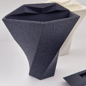 May include: A dark gray vase with a textured, faceted surface and a wide, angular opening. Behind it sits a similar white vase, and in the lower right corner is a small, smooth, dark gray rectangular component with a central cutout.