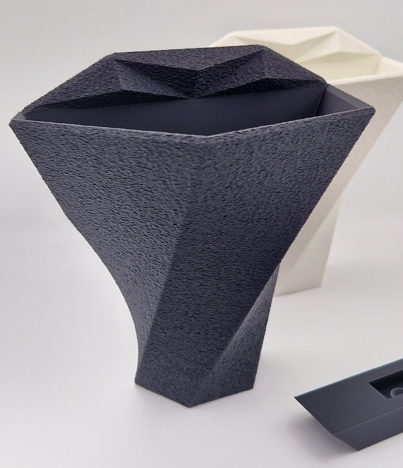 May include: A dark gray vase with a textured, faceted surface and a wide, angular opening. Behind it sits a similar white vase, and in the lower right corner is a small, smooth, dark gray rectangular component with a central cutout.