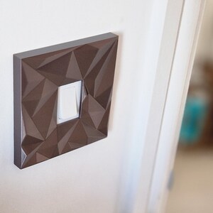 May include: A square light switch cover plate featuring a dark brown finish and a faceted, geometric surface texture. The cover plate surrounds a single, centered white rectangular rocker light switch, mounted flush against a plain white wall.