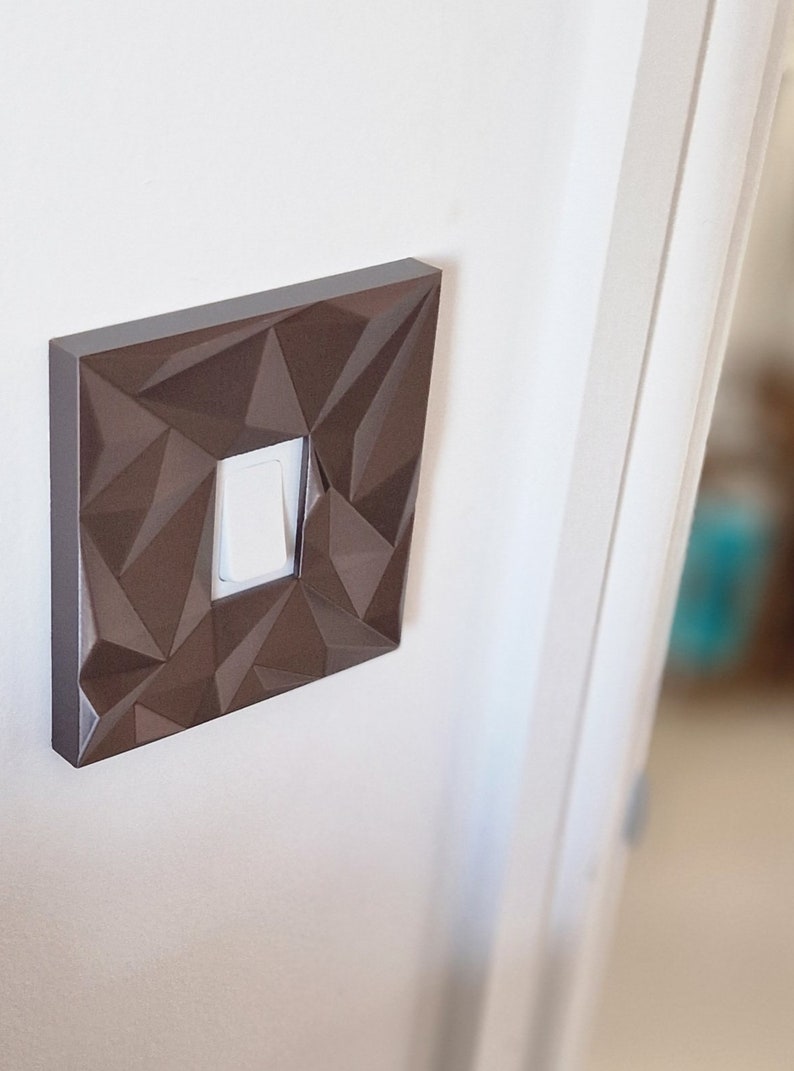 May include: A square light switch cover plate featuring a dark brown finish and a faceted, geometric surface texture. The cover plate surrounds a single, centered white rectangular rocker light switch, mounted flush against a plain white wall.