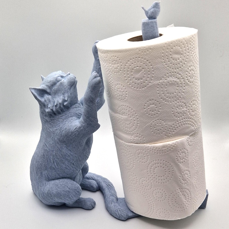 May include: A light blue ceramic cat figurine holding a roll of white paper towels. The cat is sitting on its hind legs with its front paws reaching up to hold the paper towel roll. The cat has a small bird perched on its head.