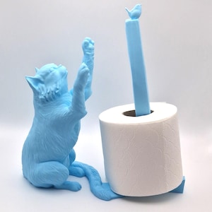 May include: A light blue cat figurine holding a roll of toilet paper. The cat is sitting on a light blue base with a bird on top of the roll holder.