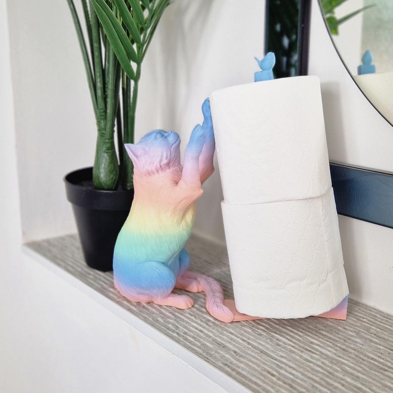 May include: A rainbow-colored cat figurine holding a roll of white toilet paper. The cat is standing on a white surface.