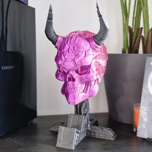 May include: A pink metallic skull with horns on a grey stand. The skull has a detailed design and appears to be made of plastic.