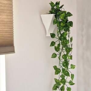 May include: A white geometric wall-mounted planter containing a trailing artificial plant with variegated green and yellow heart-shaped leaves. The planter has a tapered, angular form and is attached to a flat vertical surface.