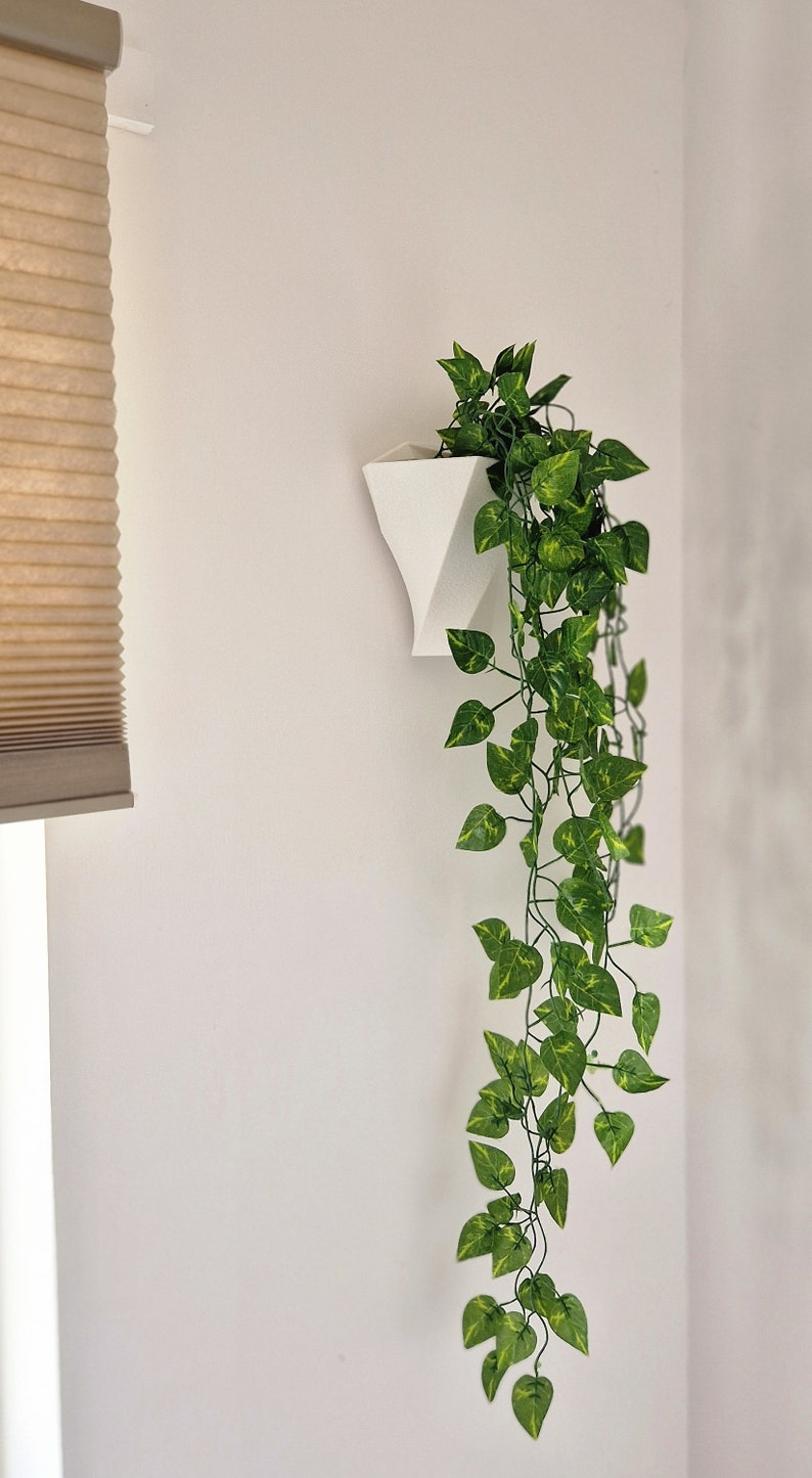 May include: A white geometric wall-mounted planter containing a trailing artificial plant with variegated green and yellow heart-shaped leaves. The planter has a tapered, angular form and is attached to a flat vertical surface.