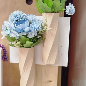 May include: Two wooden vases with a twisted design, each holding a bouquet of light blue artificial flowers. The vases are mounted on a white board.