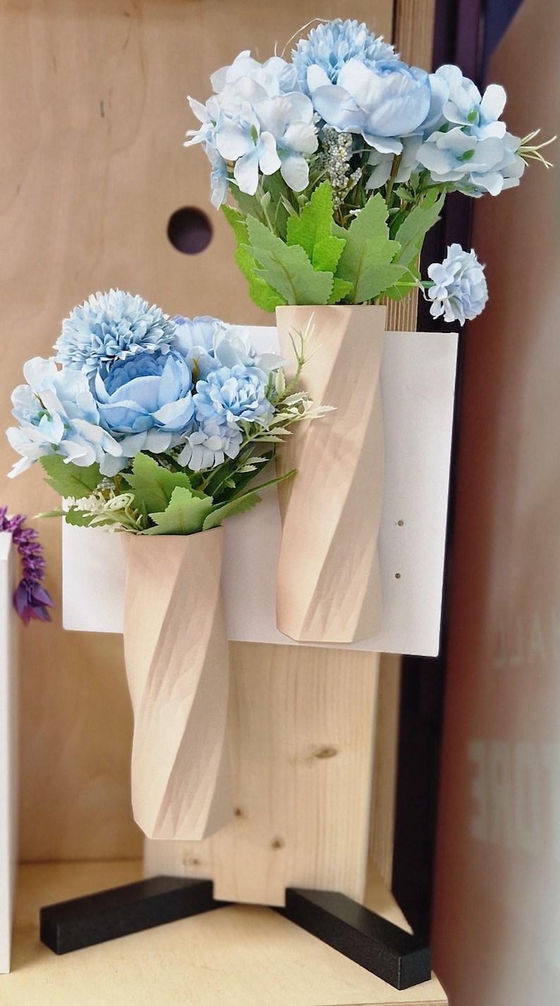 May include: Two wooden vases with a twisted design, each holding a bouquet of light blue artificial flowers. The vases are mounted on a white board.