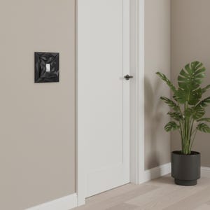 May include: A black geometric light switch cover with a white switch. The cover has a textured, angular design. The switch is mounted on a beige wall next to a white door and a potted plant.