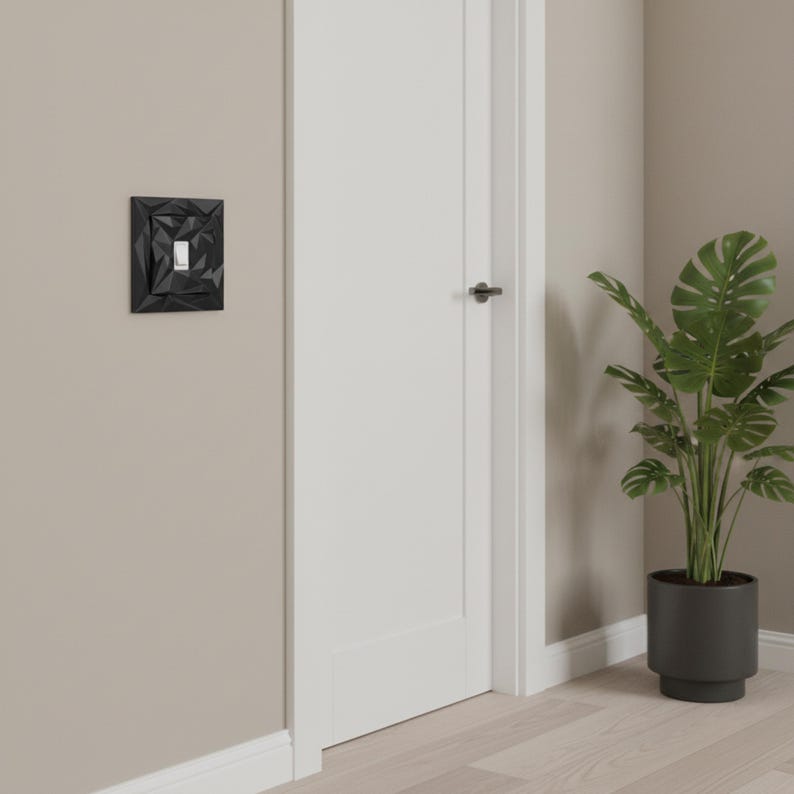 May include: A black geometric light switch cover with a white switch. The cover has a textured, angular design. The switch is mounted on a beige wall next to a white door and a potted plant.