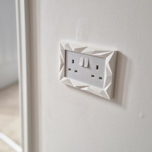 May include: White electrical outlet with two switches and three plugs, framed in a white geometric design.