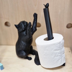 May include: A black cat figurine holding a toilet paper roll with a small black bird perched on top. The toilet paper roll is white with a patterned design.