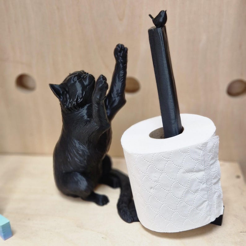 May include: A black cat figurine holding a toilet paper roll with a small black bird perched on top. The toilet paper roll is white with a patterned design.