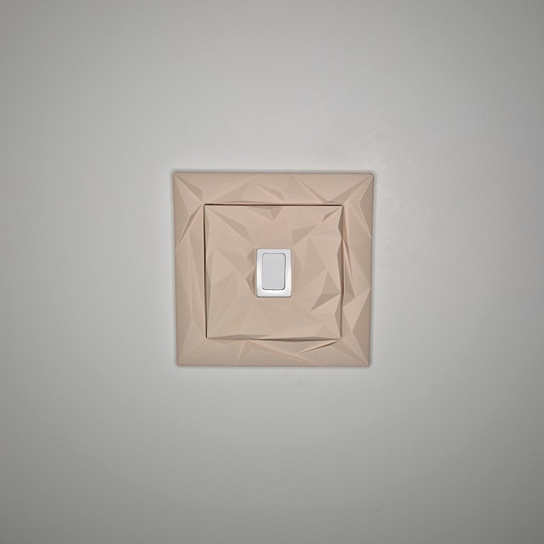 May include: A light beige, geometric-patterned light switch cover with a rectangular, silver-colored switch. The cover has a square shape with a textured, folded paper-like design. The switch is centered within the cover.