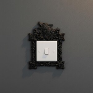 May include: A decorative light switch cover with a black, ornate frame featuring a wolf's head at the top. The white switch is centered within the frame, mounted on a gray wall. The frame has floral and scrollwork details.