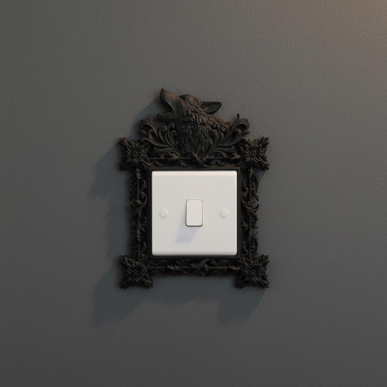 May include: A decorative light switch cover with a black, ornate frame featuring a wolf's head at the top. The white switch is centered within the frame, mounted on a gray wall. The frame has floral and scrollwork details.