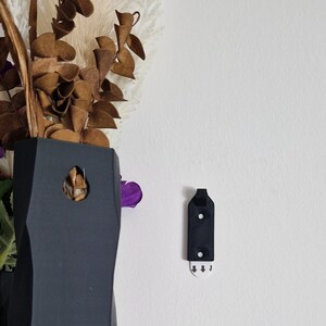 May include: A black plastic wall hook with two holes and a small white sticker with arrows pointing down. The hook is designed to hold a vase or other decorative items.