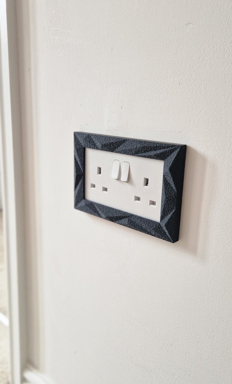 May include: A white electrical outlet and light switch with two toggles, framed in a black geometric design.