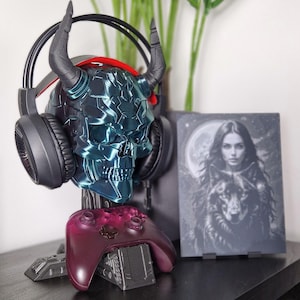 May include: A teal blue skull-shaped headset stand with horns, holding a black headset and a purple video game controller. The stand is on a black surface next to a framed black and white print of a woman with a wolf.