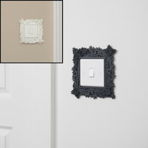 May include: Decorative light switch cover with an ornate floral design. The cover is square and surrounds a white light switch. The frame is a dark gray color, adding a touch of elegance to the wall.