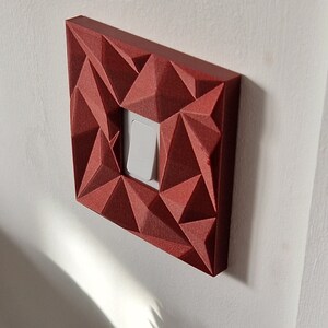 May include: A square light switch cover plate featuring a dark red, faceted, geometric, three-dimensional surface texture. The cover plate surrounds a single, white, rectangular rocker-style light switch mounted on a flat, off-white wall surface.