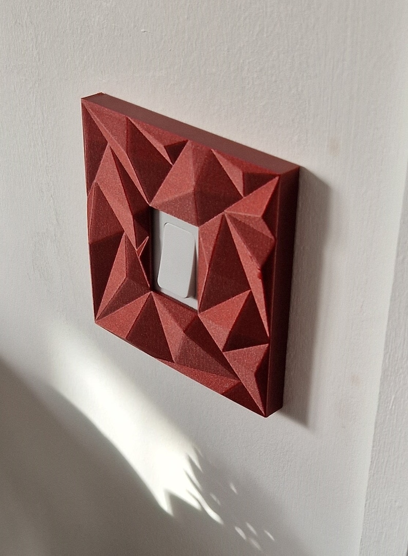 May include: A square light switch cover plate featuring a dark red, faceted, geometric, three-dimensional surface texture. The cover plate surrounds a single, white, rectangular rocker-style light switch mounted on a flat, off-white wall surface.