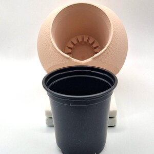 May include: A beige ceramic planter with a round shape and a hole in the top. A black plastic pot is placed inside the ceramic planter.