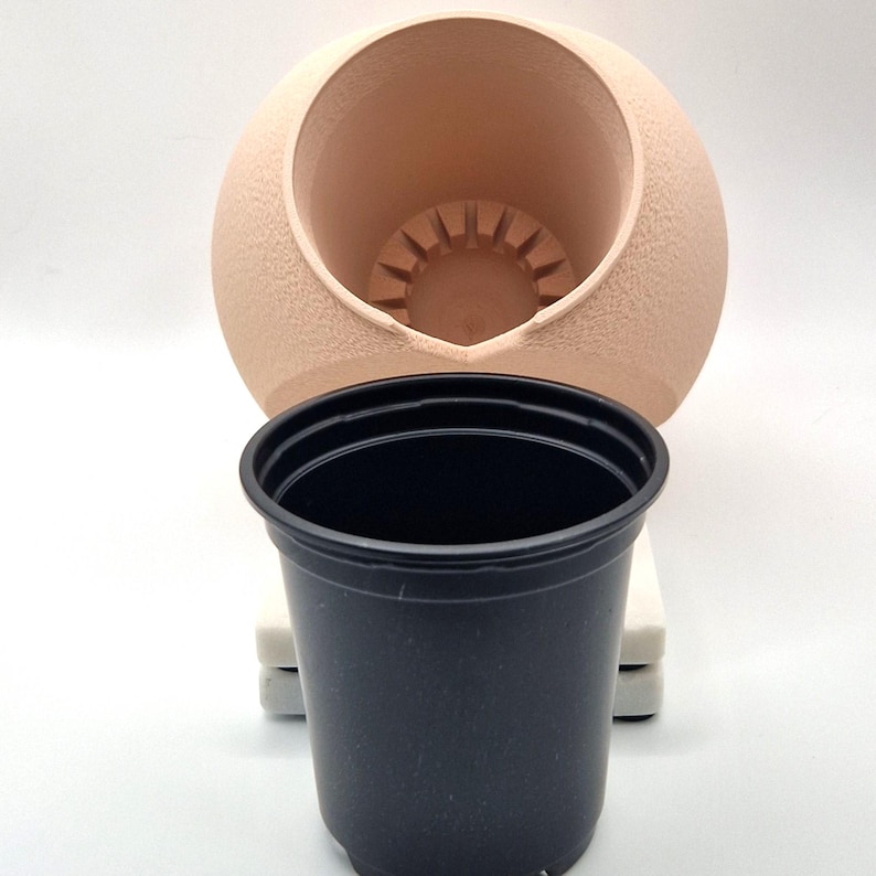 May include: A beige ceramic planter with a round shape and a hole in the top. A black plastic pot is placed inside the ceramic planter.