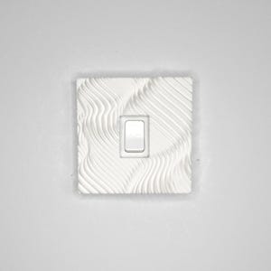 May include: A square white light switch cover plate featuring a textured surface with raised, flowing, wave-like lines. The plate has a centered rectangular cutout for a single rocker-style light switch, which is installed in the middle of the plate.