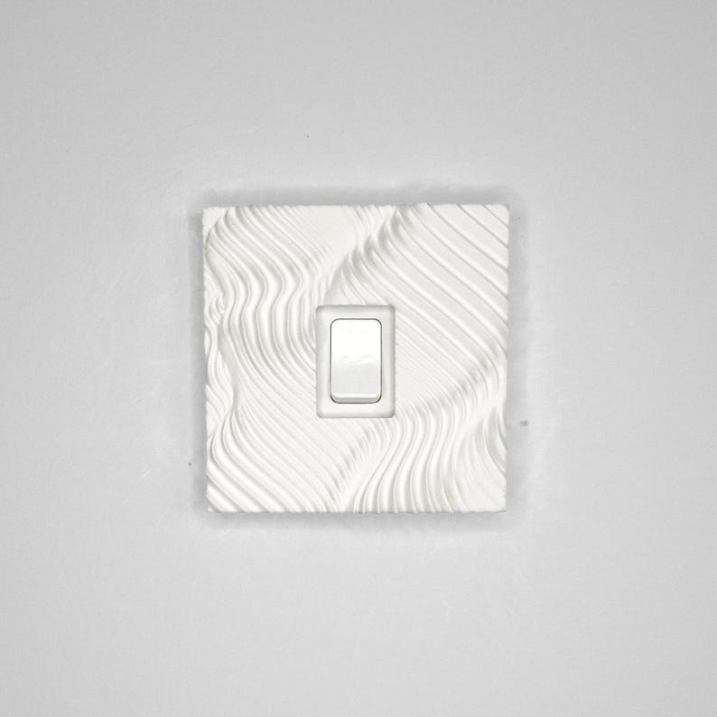 May include: A square white light switch cover plate featuring a textured surface with raised, flowing, wave-like lines. The plate has a centered rectangular cutout for a single rocker-style light switch, which is installed in the middle of the plate.
