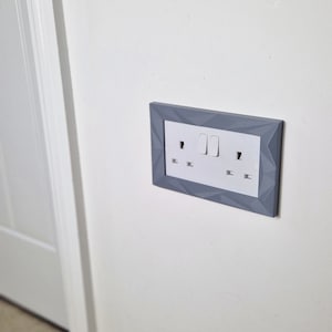 May include: A white electrical outlet and light switch with two toggle switches, framed in a gray geometric design.