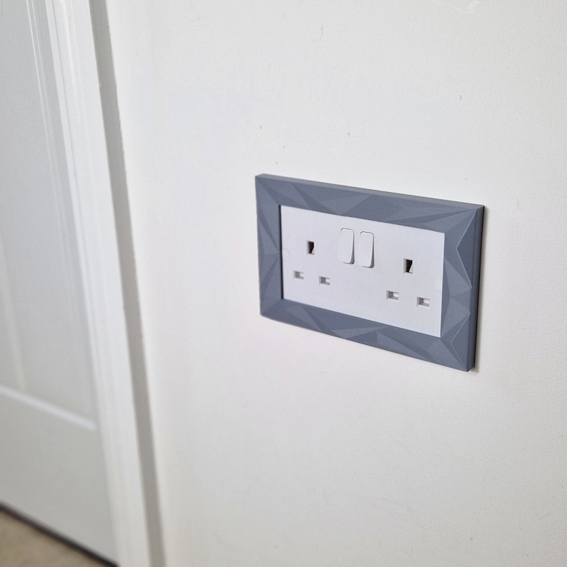 May include: A white electrical outlet and light switch with two toggle switches, framed in a gray geometric design.