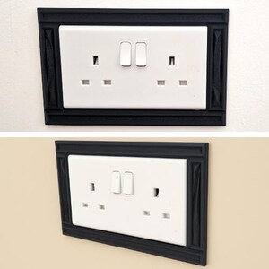 May include: A white double electrical wall outlet with two switches, framed by a rectangular black decorative cover plate. The frame features a textured, molded border design that surrounds the standard rectangular white plastic socket unit.