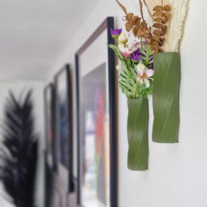 May include: Two green, textured, cylindrical vases with a twist design are mounted on a white wall. The vases hold a bouquet of purple and white flowers.