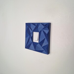 May include: A square light switch cover plate featuring a dark blue, faceted geometric surface texture. The cover plate has a central rectangular cutout that frames a standard white rocker-style light switch toggle centered within the design.