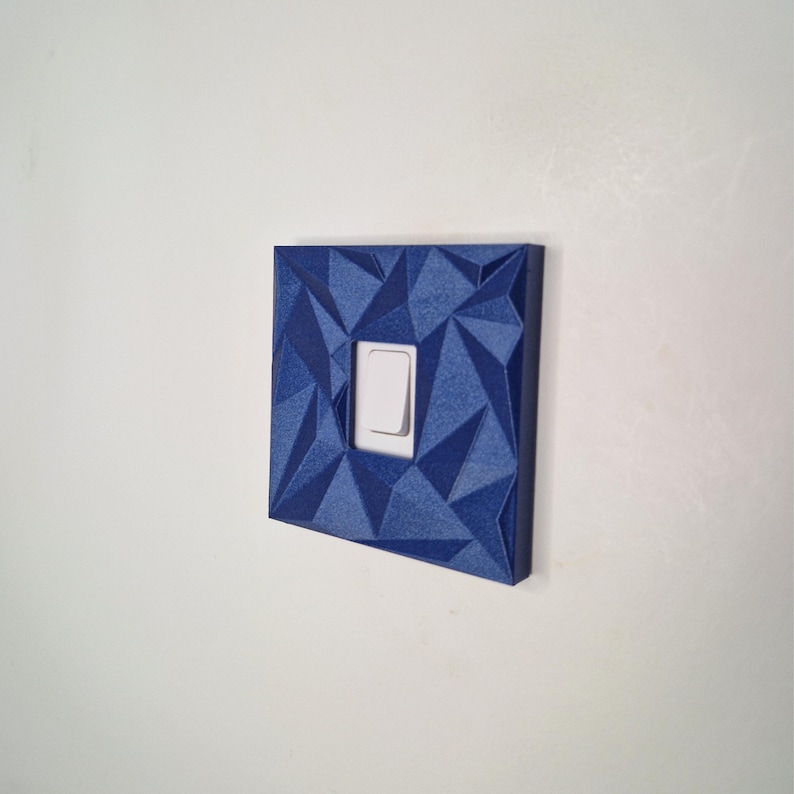 May include: A square light switch cover plate featuring a dark blue, faceted geometric surface texture. The cover plate has a central rectangular cutout that frames a standard white rocker-style light switch toggle centered within the design.