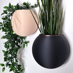 Puede incluir: Dos macetas de pared redondas, una es beige y la otra es negra. Ambas macetas tienen una superficie texturizada y están llenas de plantas verdes.