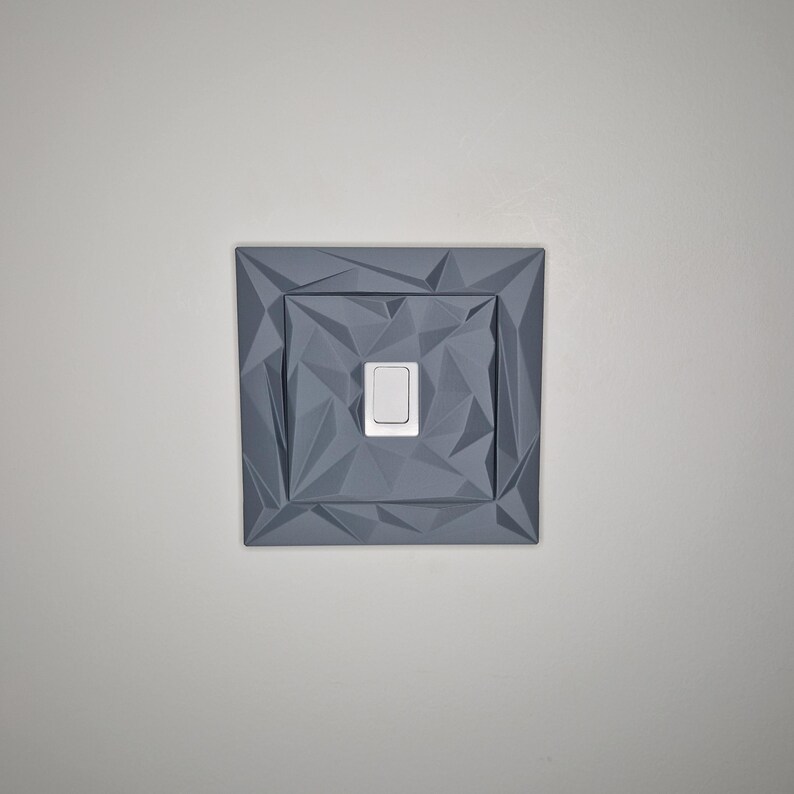 May include: A gray, geometric-patterned light switch cover with a white switch. The cover has a square shape with a textured, faceted design. The switch is rectangular and centered within the cover.