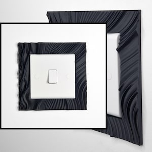 May include: A white light switch featuring a decorative black frame with a textured, wavy, and rippled surface pattern. The frame surrounds the square switch plate, which includes a central toggle and two circular screw covers on the sides.