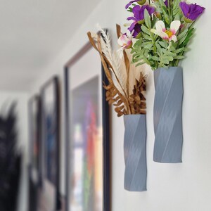 May include: Two grey, textured, wall-mounted vases with a twisted design. One vase holds a bouquet of purple and white flowers, while the other holds dried brown branches.