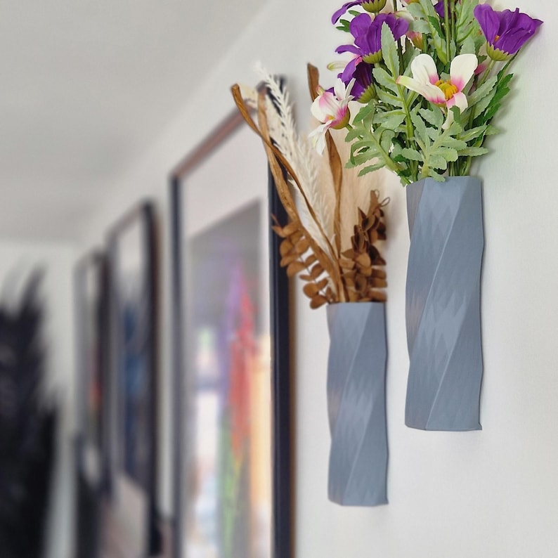 May include: Two grey, textured, wall-mounted vases with a twisted design. One vase holds a bouquet of purple and white flowers, while the other holds dried brown branches.