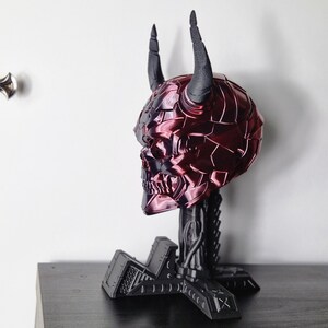 May include: A red and black skull sculpture with horns, standing on a black metal base. The skull is detailed with a textured, segmented surface.
