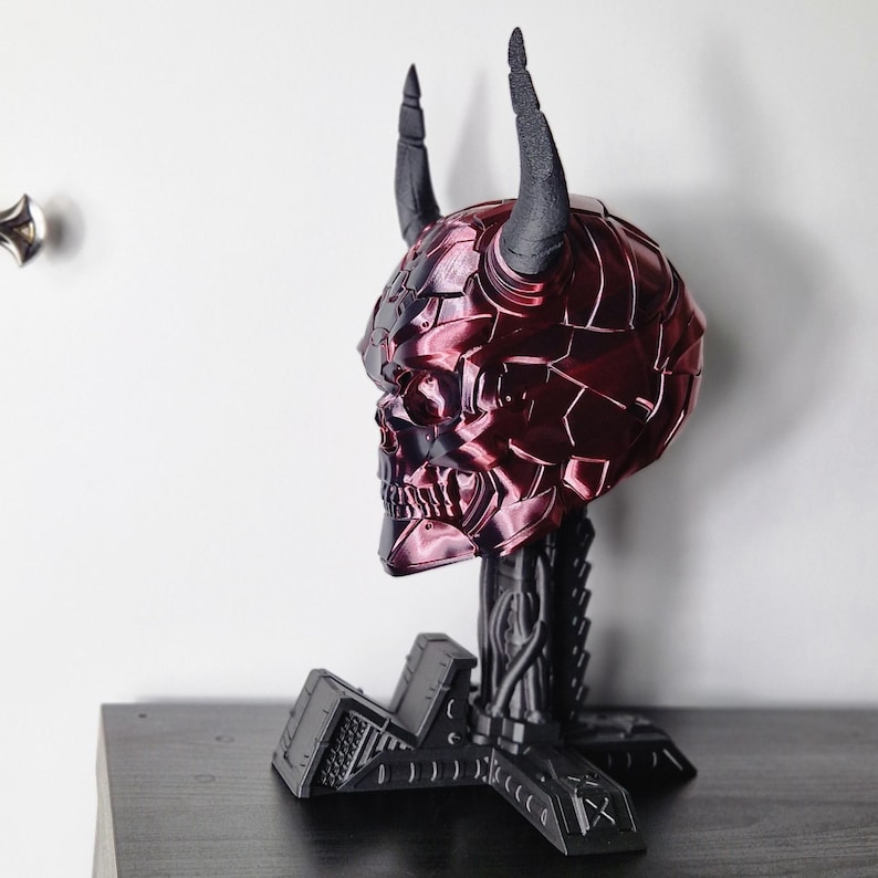 May include: A red and black skull sculpture with horns, standing on a black metal base. The skull is detailed with a textured, segmented surface.