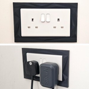 Może przedstawiać: Białe podw&oacute;jne gniazdko elektryczne z czarną geometryczną ramką o fasetowanej, kanciastej fakturze powierzchni. Gniazdko zawiera dwa przełączniki i dwa otwory, z włożoną czarną wtyczką oraz szarą wtyczką marki Shark.