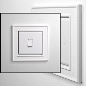 May include: A white square light switch featuring a central rocker toggle mechanism, surrounded by a decorative square frame with beveled edges. The switch plate has a smooth finish and includes two small circular screw covers on either side.