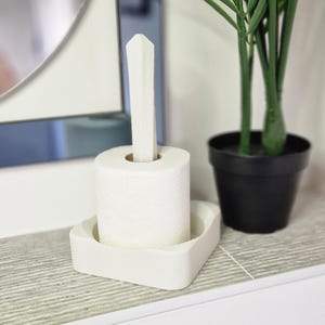 Puede incluir: Porta rollo de papel higi&eacute;nico de cer&aacute;mica blanca con un rollo de papel higi&eacute;nico blanco. El soporte tiene una base cuadrada y una parte superior alta, delgada y puntiaguda.