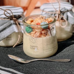 Puede incluir: Vela en tarro de cristal transparente con etiqueta Captain Crunch, rellena de cera clara y cubierta con trozos de cereales. La vela forma parte de un conjunto, con una cuchara en primer plano.
