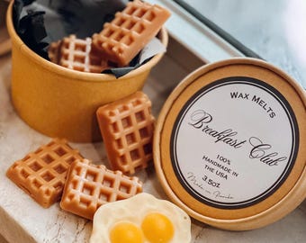 May include: A round cardboard container filled with light brown waffle-shaped wax melts, accompanied by a white egg-shaped wax melt with two yellow yolks. The lid features the text WAX MELTS Breakfast Club 100% HANDMADE IN THE USA 3.5oz.