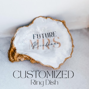 May include: A decorative oyster shell ring dish with a white interior and gold-painted edges. The text FUTURE MRS is printed in black and rose gold, with the name Riedeman written in black script across the center of the shell surface.